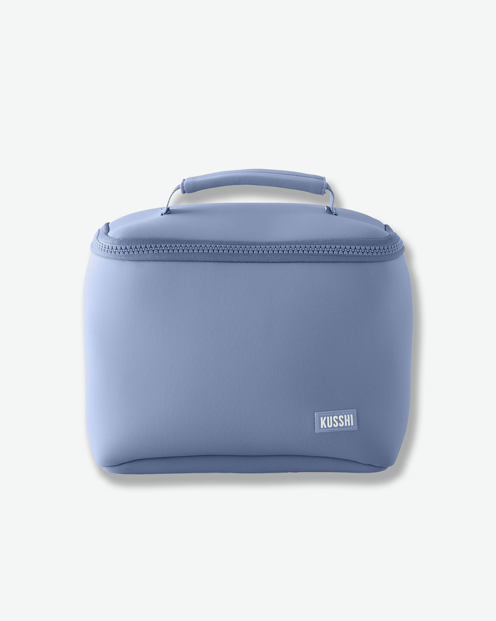 A studio product shot of a structured, periwinkle blue fabric makeup bag from Kusshi against a light gray background. The rectangular bag features rounded sides, a matching blue zipper closure that wraps around the upper edge, and a sturdy top handle. A small, rectangular white logo patch with the word 'KUSSHI' is stitched onto the bottom right corner.