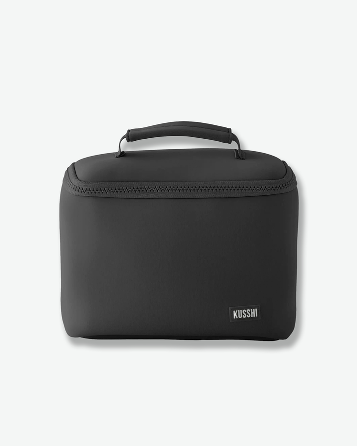 A studio product shot of a structured, matte black fabric makeup bag from Kusshi against a light gray background. The bag features a rectangular shape with curved vertical corners, a thick black top handle, and a matching black zipper along the upper lid. A small rectangular black and white logo patch reading 'KUSSHI' sits on the lower right corner.