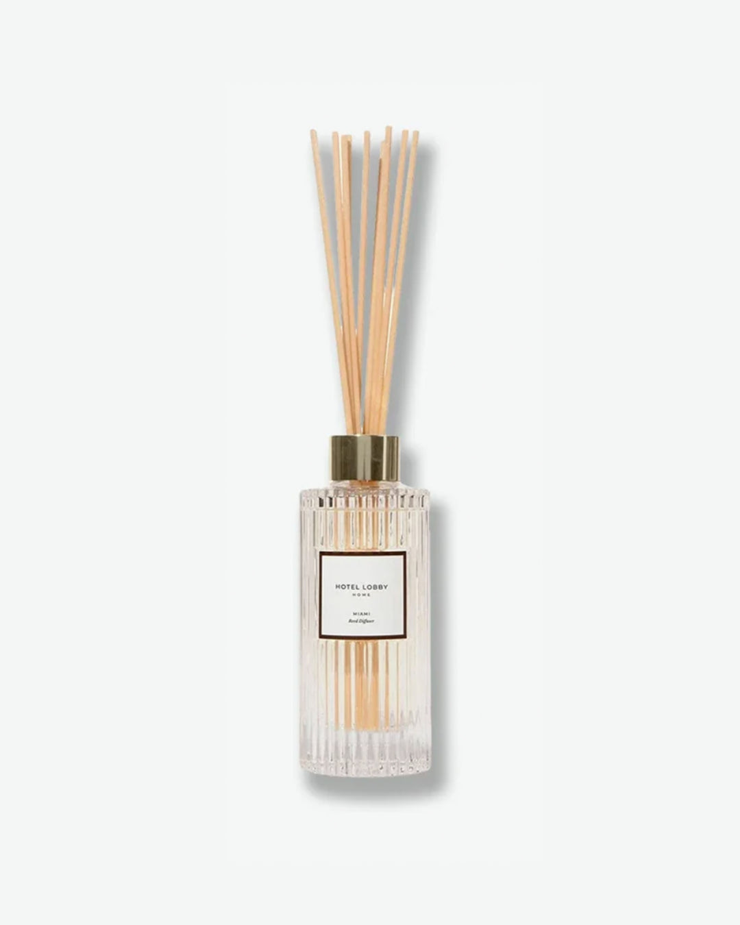 Hotel Lobby Home Miami Reed Diffuser featuring a fluted clear glass bottle filled with clear liquid and light wooden reeds extending upward, isolated against a light grey background.
