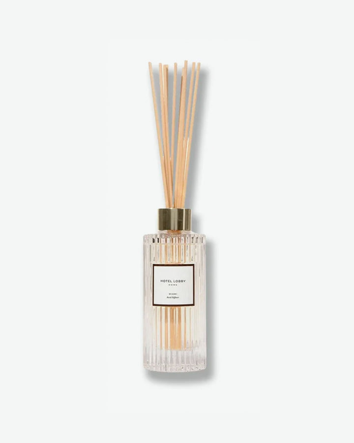 Hotel Lobby Home Miami Reed Diffuser featuring a fluted clear glass bottle filled with clear liquid and light wooden reeds extending upward, isolated against a light grey background.