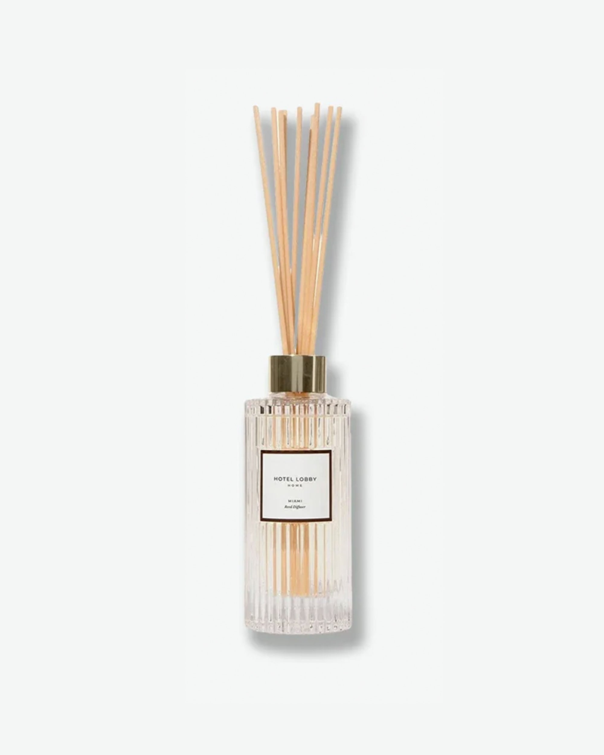 Hotel Lobby Home Miami Reed Diffuser featuring a fluted clear glass bottle filled with clear liquid and light wooden reeds extending upward, isolated against a light grey background.