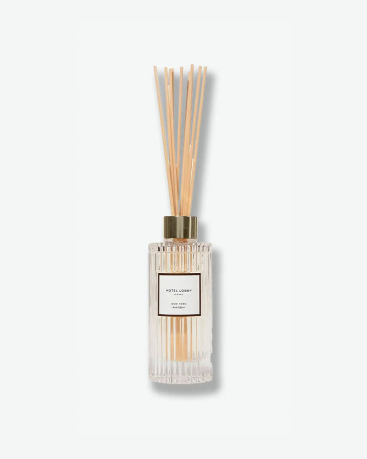 Hotel Lobby Home New York Reed Diffuser featuring a fluted clear glass bottle filled with clear liquid and light wooden reeds extending upward, isolated against a light grey background.