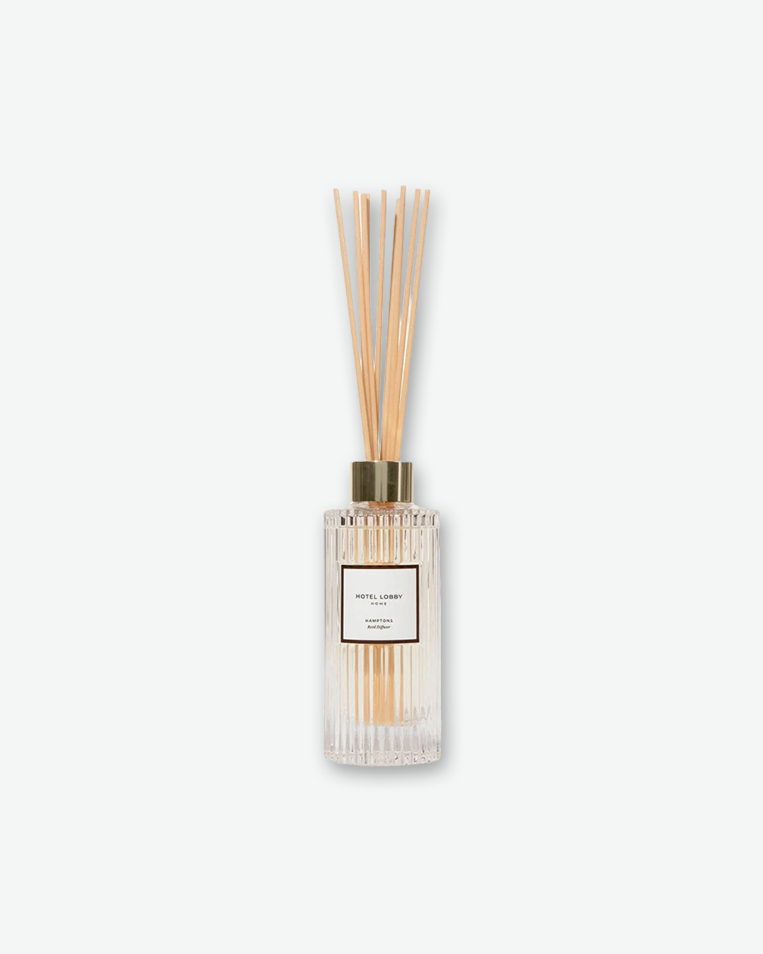 A studio product shot of a Hotel Lobby Home reed diffuser against a light gray background. The diffuser features a clear glass bottle with a fluted, ribbed texture and a square white label that reads 'HOTEL LOBBY HOME' and 'HAMPTONS Reed Diffuser'. A metallic gold collar sits at the neck of the bottle, holding a cluster of natural, light wood diffuser reeds fanning upward.