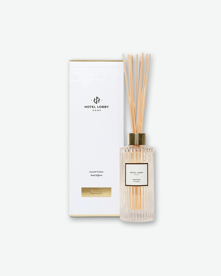 A product presentation shot showing the Hotel Lobby Home reed diffuser standing next to its original packaging box. The tall, rectangular box is white with a minimalist layout, displaying the brand's interlocking monogram logo and text reading 'HOTEL LOBBY HOME' near the top and 'HAMPTONS Reed Diffuser' near the bottom. A small gold metallic rectangle accents the base of the box, matching the gold collar on the glass diffuser jar beside it.