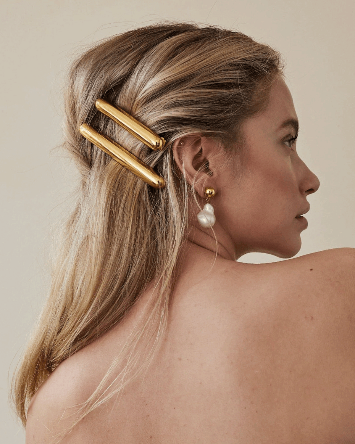 A woman shown from the back and side wearing a gold hair claw clip holding her hair in place, with a pearl drop earring also visible against a plain beige background.
