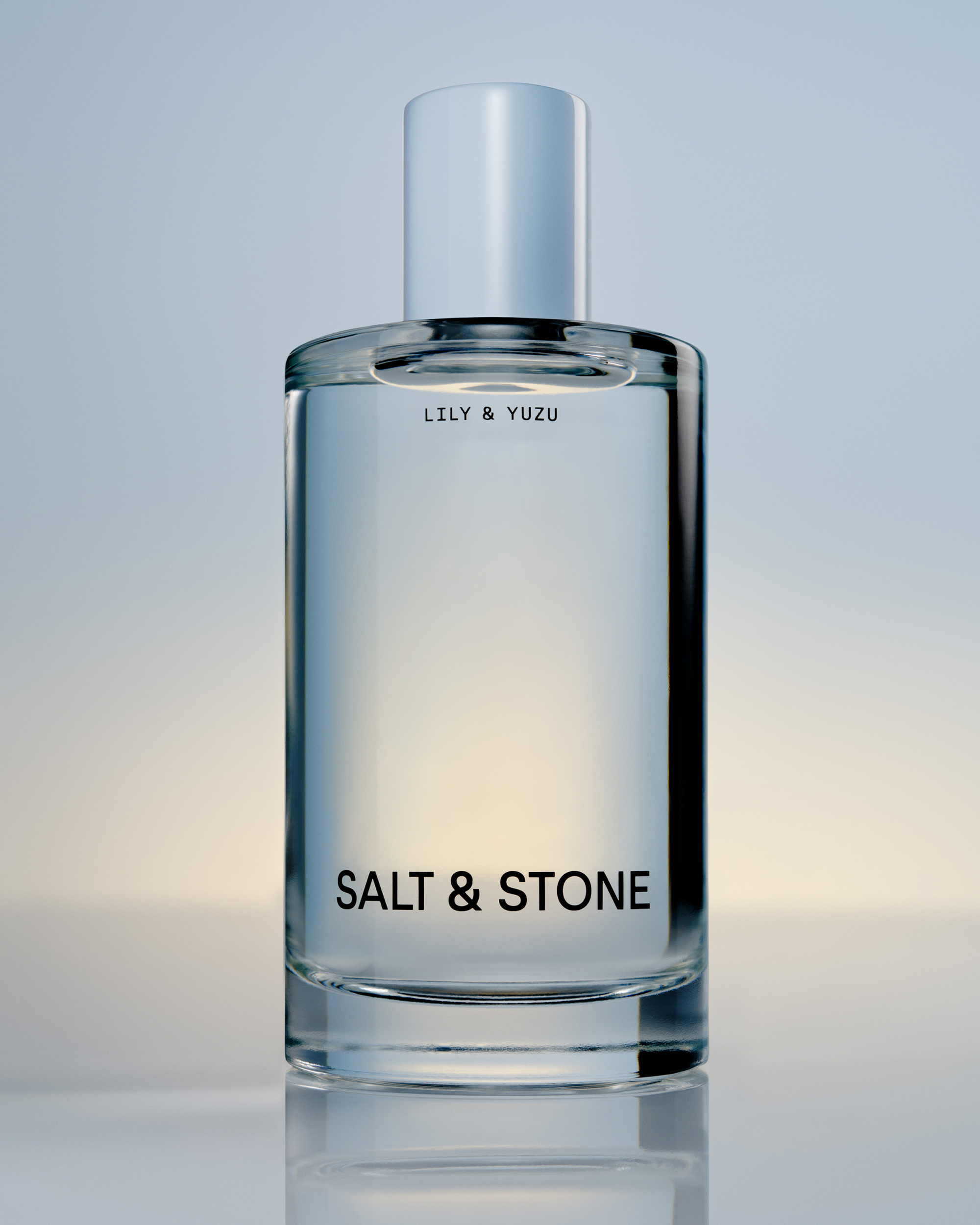 A close-up of a clear glass bottle of Salt & Stone "Lily & Yuzu" fragrance against a soft, gradient background.