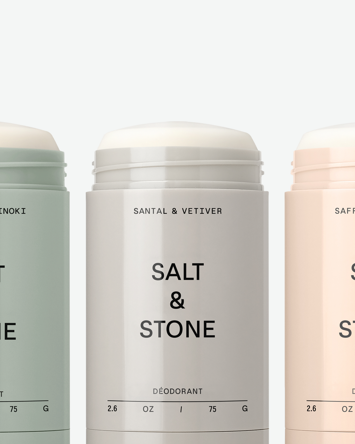 Close-up of three un-capped Salt & Stone deodorant sticks in sage green, grey, and blush pink against a plain background.