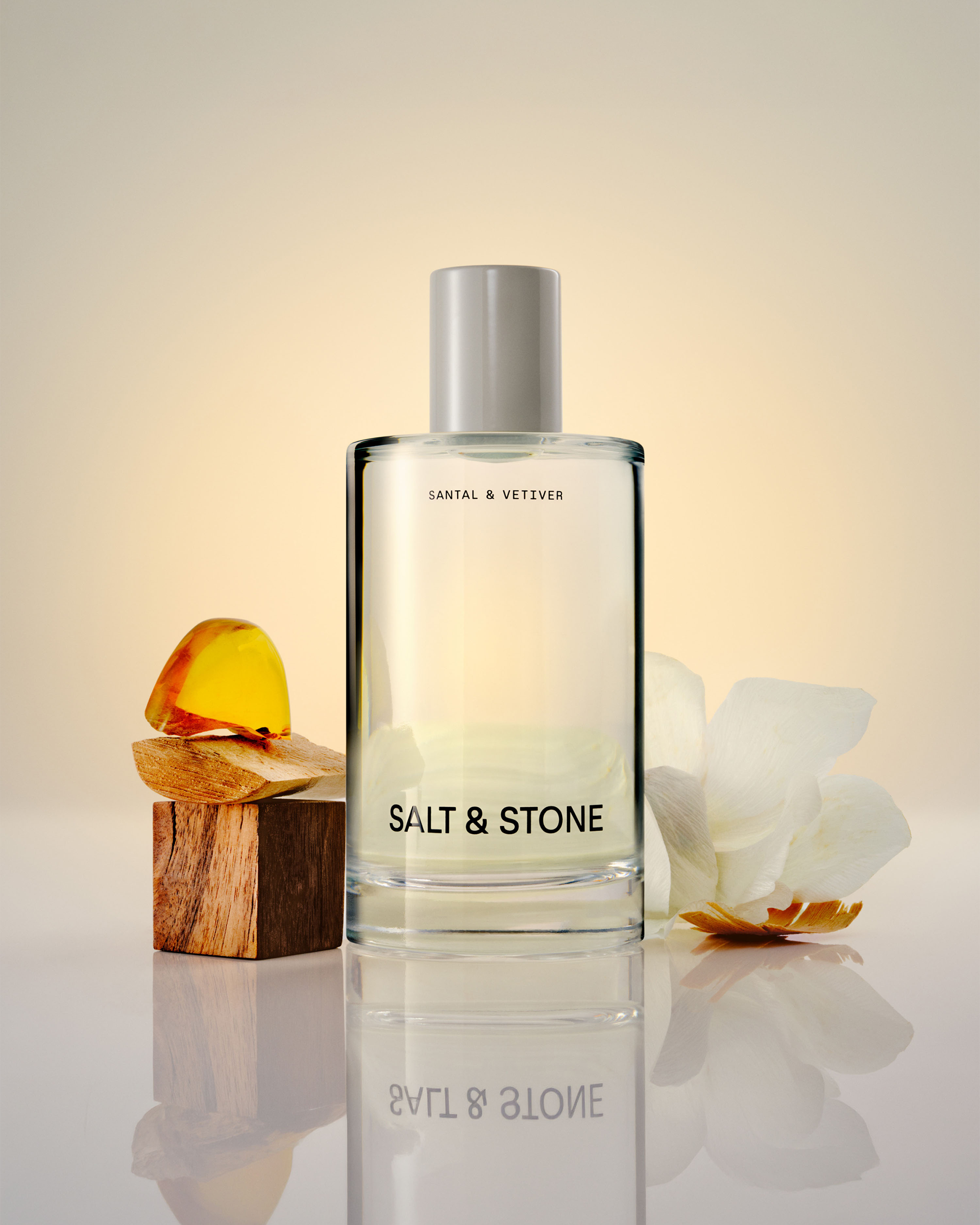 A clear rectangular perfume bottle labeled 'SALT & STONE' by Santal & Vetiver, with a matte silver cap, set on a reflective surface. A small wooden block with amber pieces and a white flower sit beside it against a warm cream background.
