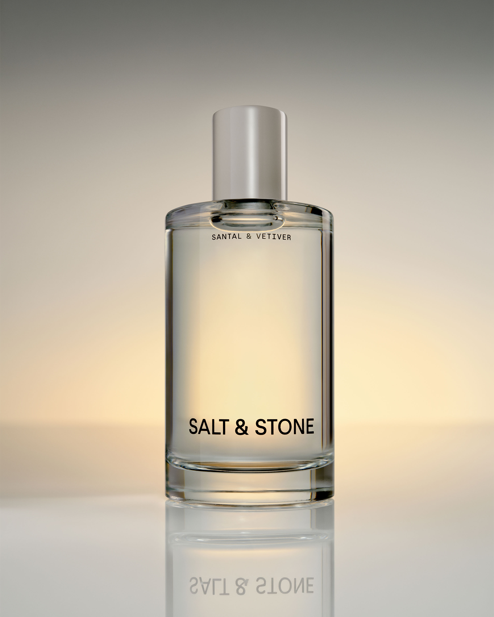 A clear glass bottle of Santal & Vetiver Salt & Stone perfume with a silver cap, centered on a reflective surface against a warm beige and gray gradient background.