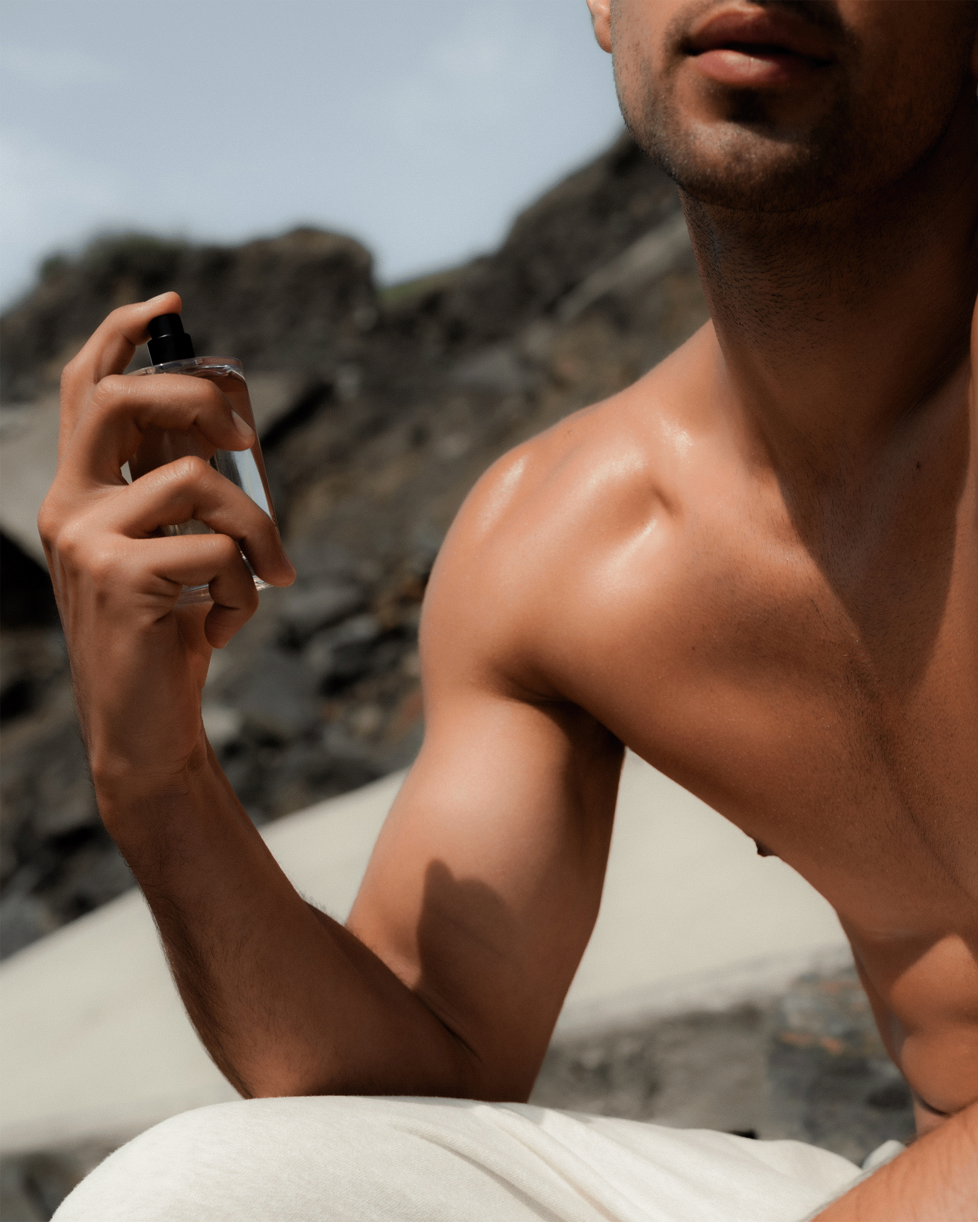 A close-up lifestyle shot of a man outdoors on a rocky beach or mountain, holding and spraying a clear bottle of fragrance. He is shirtless, wearing white pants, with natural light highlighting his skin and the bottle.