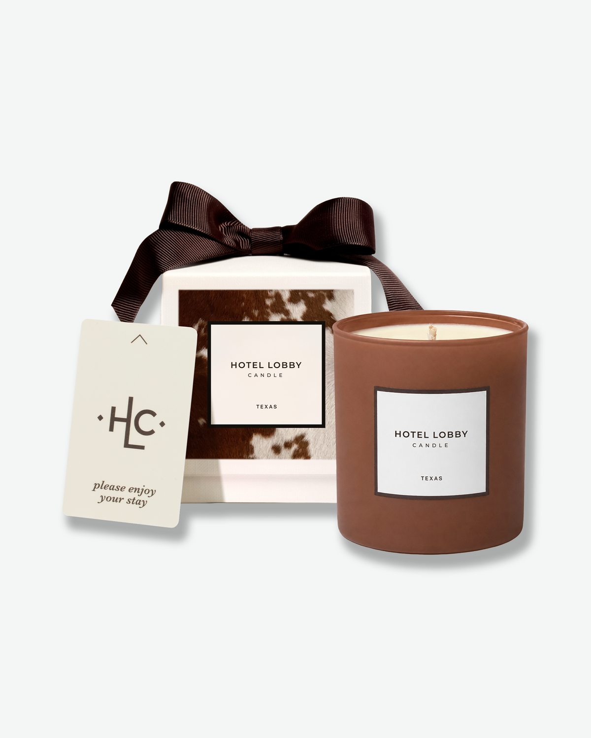 Hotel Lobby Candle Texas candle displayed with its cowhide-inspired gift box, brown ribbon, and “please enjoy your stay” card.
