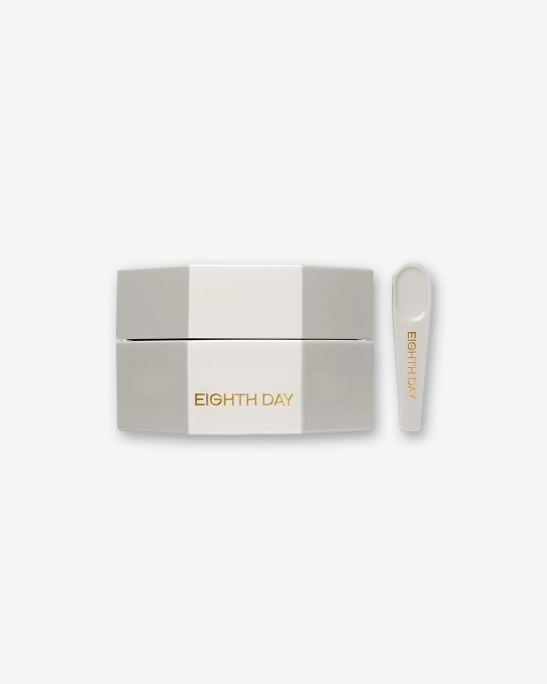 A product shot of an Eighth Day skincare jar and applicator tool, centered against a plain light gray background. The jar is a distinct, light gray octagonal shape with the words "EIGHTH DAY" printed in gold on the front side. Standing upright to its right is a matching light gray plastic spatula tool with "EIGHTH DAY" written in gold down the handle.