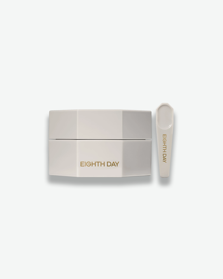 A product shot of an Eighth Day skincare jar and applicator tool, centered against a plain light gray background. The jar is a distinct, light gray octagonal shape with the words "EIGHTH DAY" printed in gold on the front side. Standing upright to its right is a matching light gray plastic spatula tool with "EIGHTH DAY" written in gold down the handle.