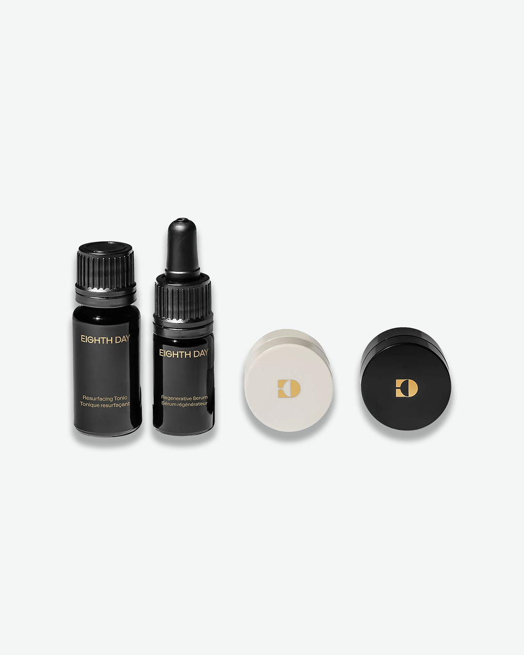 A product shot showing the individual components of the Eighth Day travel skincare kit on a light gray background. Arranged in a row from left to right are a miniature Resurfacing Tonic bottle, a miniature Regenerative Serum dropper bottle, a small white jar lid with a gold logo, and a small black jar lid with a gold logo.