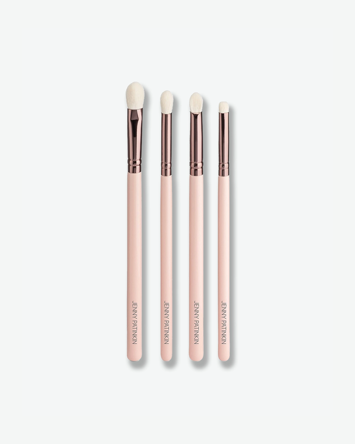 A product shot of four slender eye makeup brushes arranged vertically against a plain light gray background. Each brush has a light pink handle with "JENNY PATINKIN" stamped in gold at the bottom, a metallic rose-gold ferrule, and fine white bristles tailored for detailing, blending, and shading.