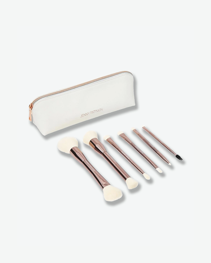 A product shot of six double-ended makeup brushes arranged on a surface in front of a slim, off-white cosmetic pouch, angled against a plain light gray background. The dual-sided brushes feature metallic rose-gold handles and soft white bristles on both ends, laid out neatly next to the textured faux-leather case which displays the name "JENNY PATINKIN" in small rose-gold lettering.