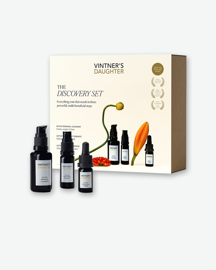 A product display shot of the Vintner's Daughter Discovery Set. In the background, a minimalist cream-colored product box features text reading 'Vintner's Daughter The Discovery Set' alongside simple floral illustrations of a poppy stem and a single orange petal. In the foreground, three small travel-sized amber glass bottles with black pumps or droppers are arranged, labeled as the Active Renewal Cleanser, Active Treatment Essence, and Active Botanical Serum.