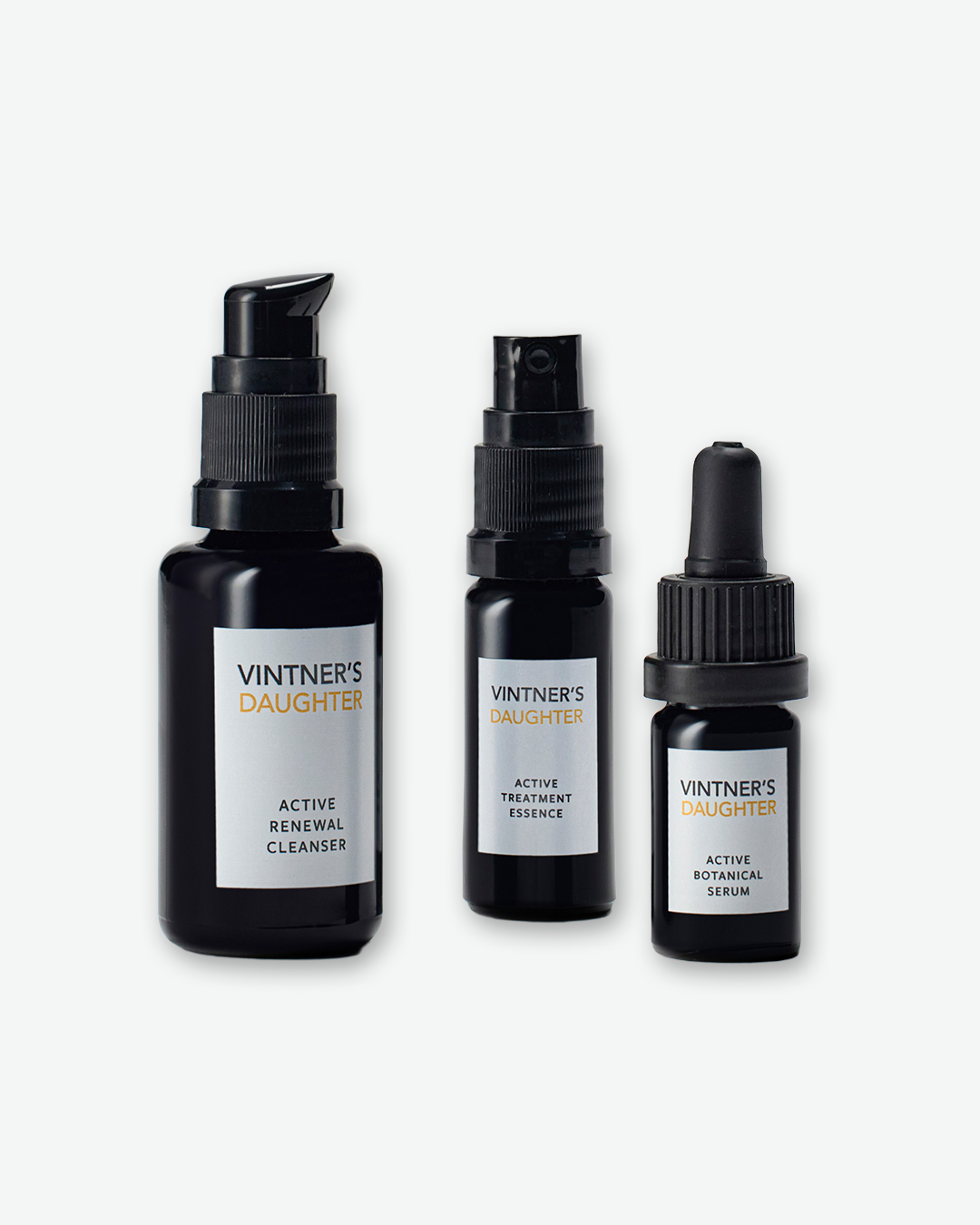 A close-up view of three travel-sized Vintner’s Daughter skincare bottles against a plain light gray background. From left to right, the dark amber bottles feature clean white labels identifying them as the Active Renewal Cleanser with a pump top, the Active Treatment Essence with a spray nozzle, and the Active Botanical Serum with a glass dropper top.