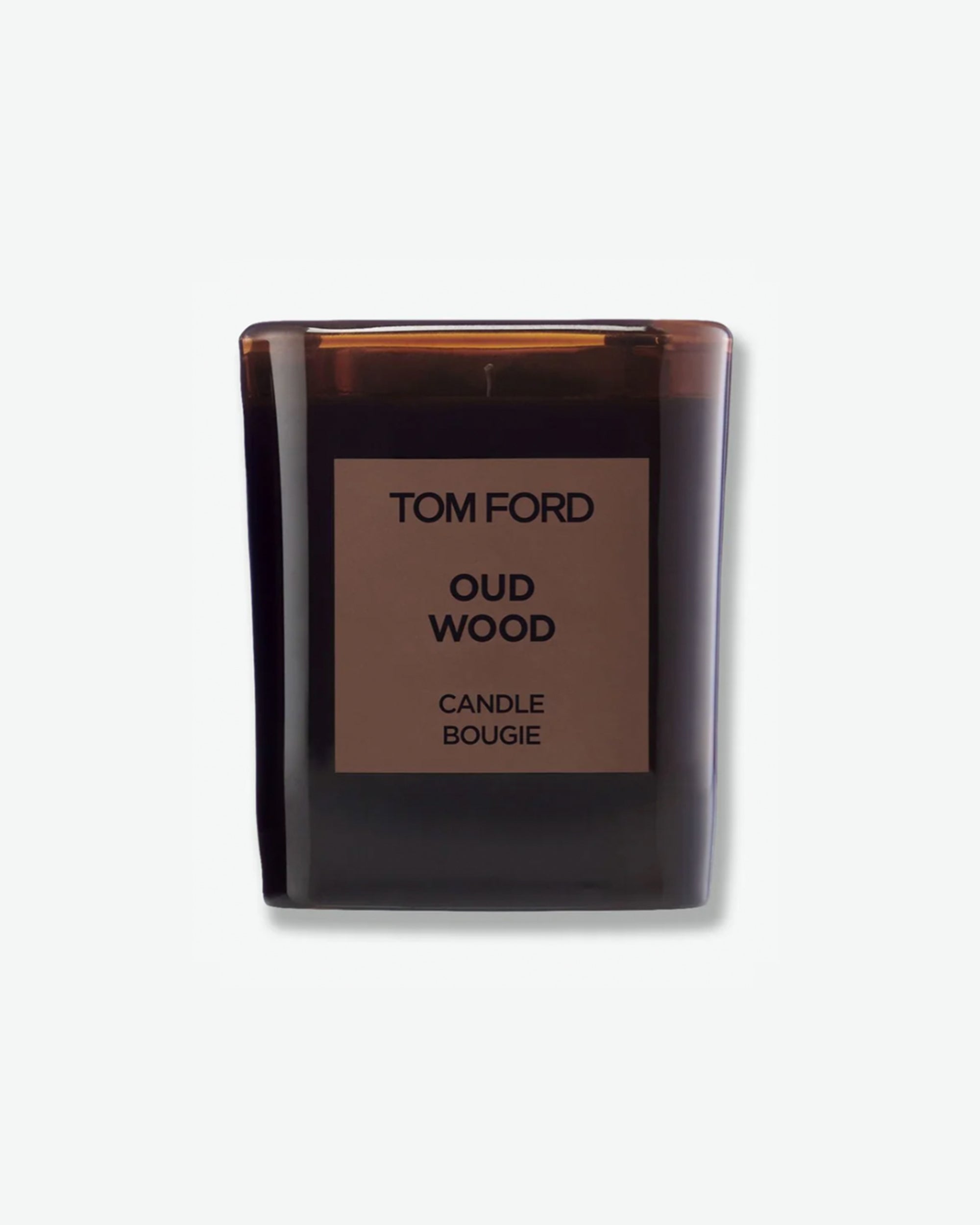 Product shot of a Tom Ford Oud Wood scented candle. The candle is contained in a glossy, translucent dark smoked-brown or amber glass jar. Centered on the front is a square muted-brown label featuring black text that reads "TOM FORD OUD WOOD CANDLE BOUGIE". The item casts a soft shadow against a clean, minimal light grey background.