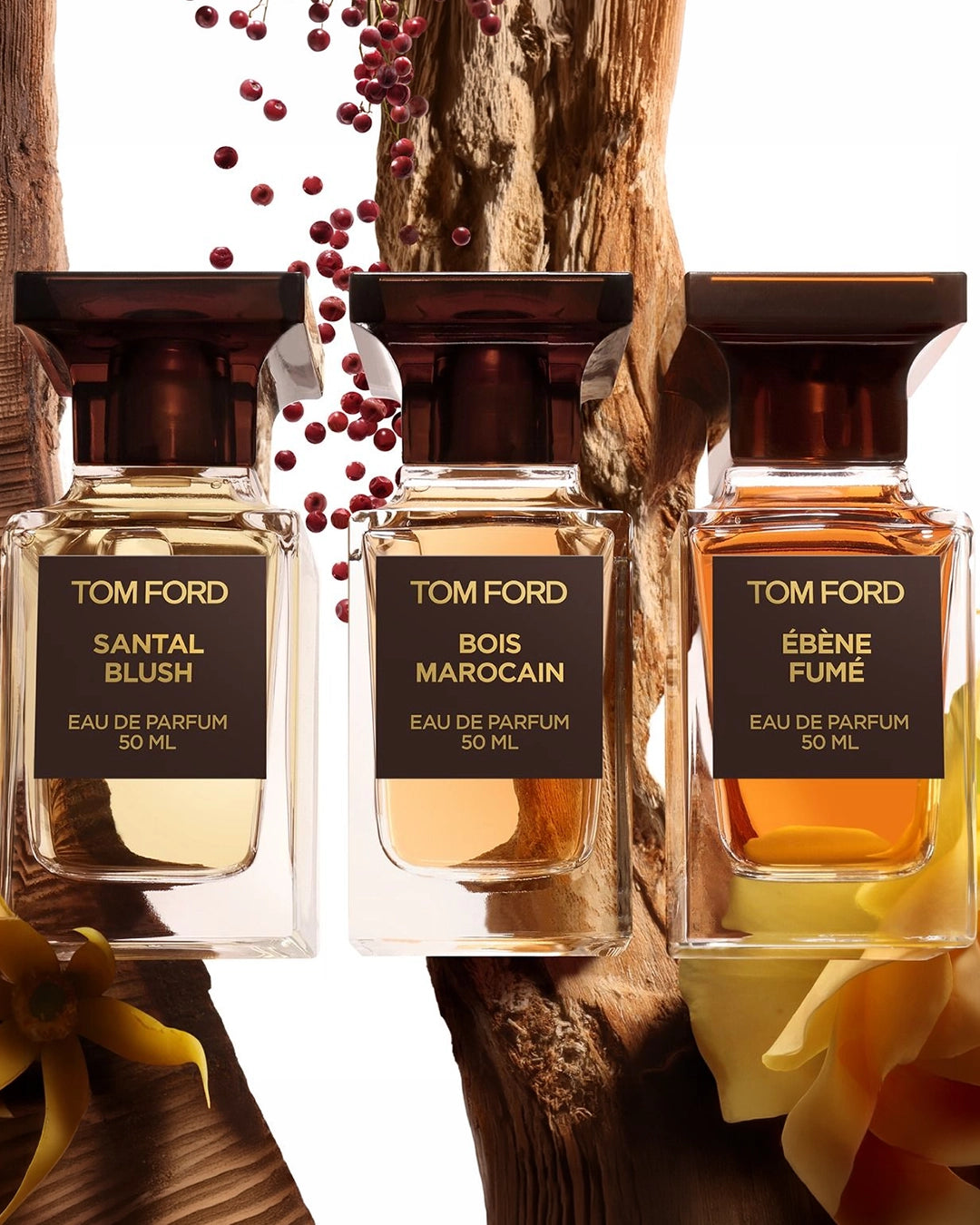 A group display of three Tom Ford Eau de Parfum bottles (50ml each) nestled against a rustic, vertical log background. From left to right: Santal Blush, Bois Marocain, and Ébène Fumé, each showcasing a different shade of amber and golden liquid. A branch of small red berries cascades down from the top center, adding a pop of color to the warm, woody composition.