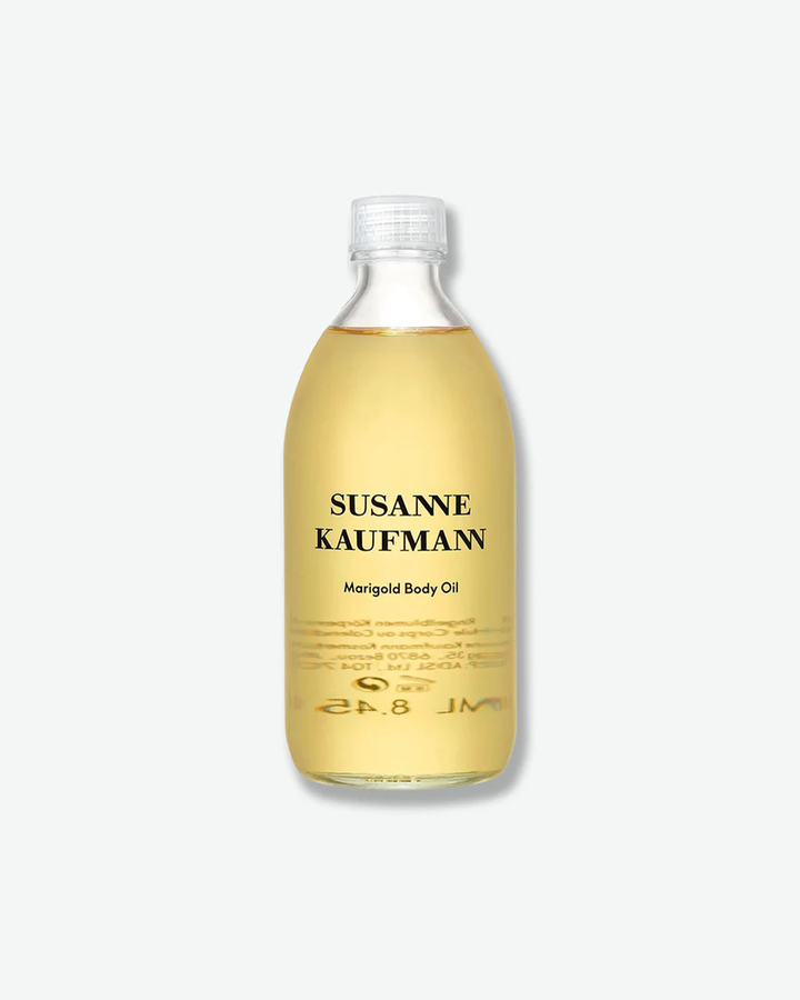 A product shot of a Susanne Kaufmann Marigold Body Oil bottle, centered against a plain light gray background. The curved clear glass bottle holds a translucent, bright golden-yellow body oil and is sealed with a clear plastic ribbed cap. The front displays clean black typography detailing the brand name and product title.