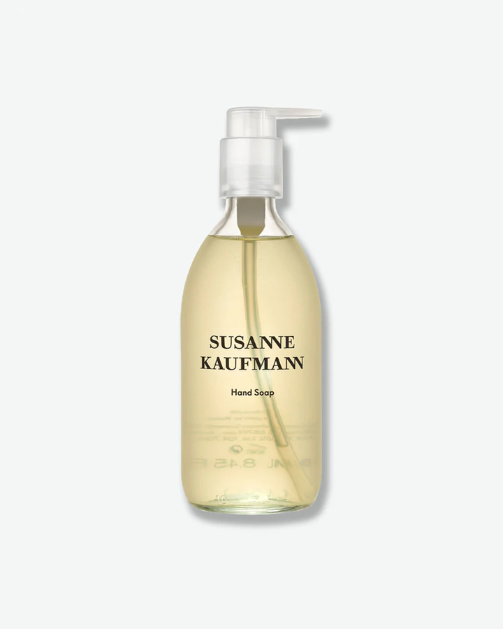 A product shot of a Susanne Kaufmann Hand Soap bottle, centered against a plain light gray background. The curved bottle is made of clear glass, revealing a translucent, light-amber liquid soap inside. It is fitted with a clear plastic pump dispenser and features black text branding on the front.
