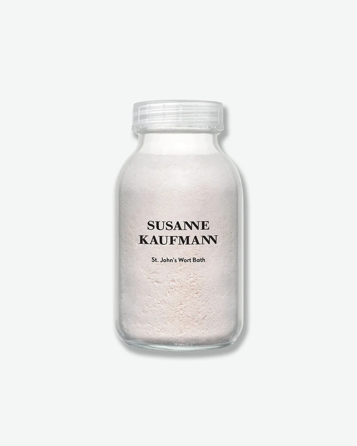 A product shot of a Susanne Kaufmann St. John's Wort Bath container, centered against a plain light gray background. The wide-mouthed clear glass jar is filled to the neck with an opaque, pale pinkish-white bath powder. It features the brand name in black serif typography printed directly on the glass above the product name, and is sealed with a clear plastic ribbed screw cap.