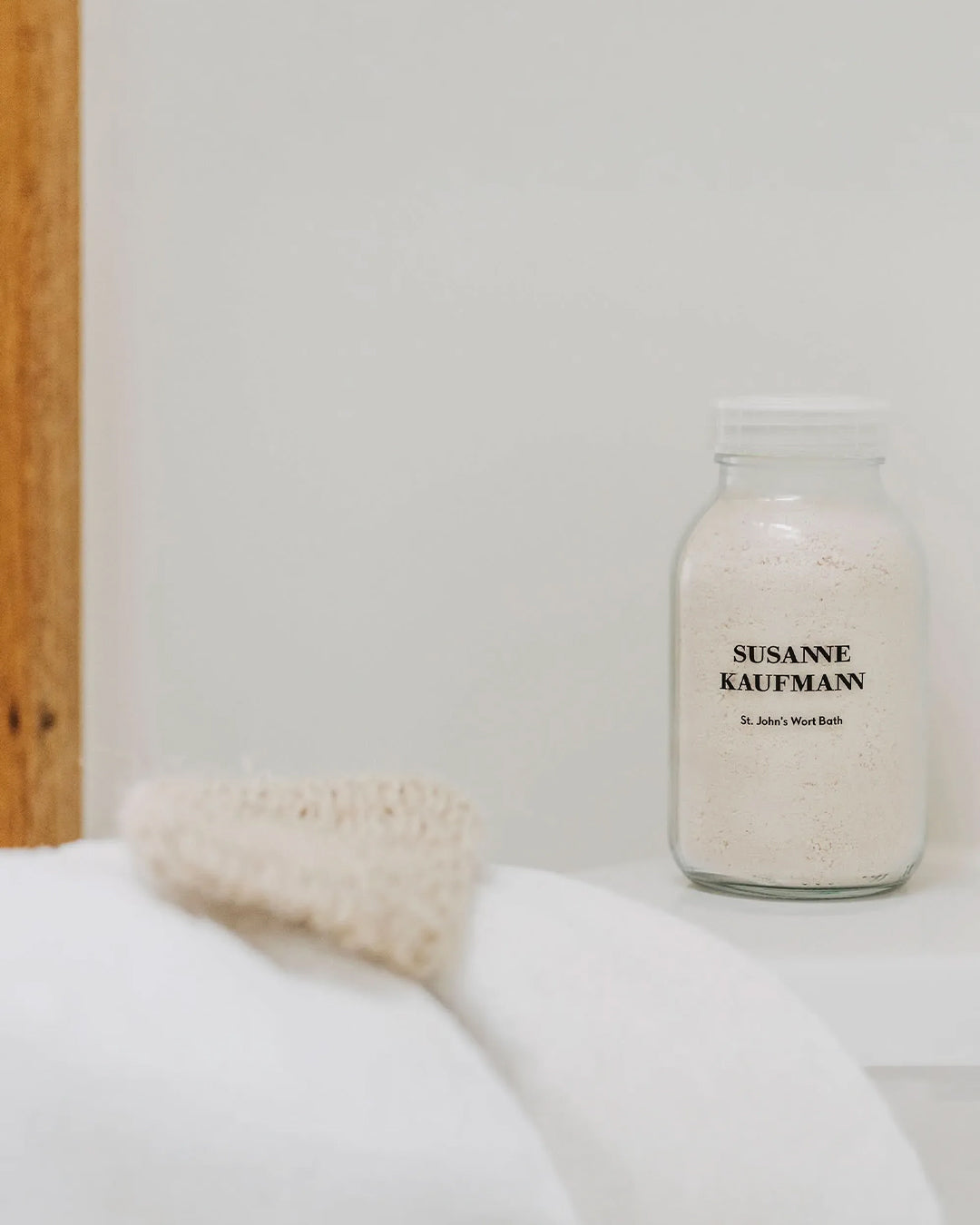 Jar of Susanne Kaufmann bath salts on a white surface with a wooden shelf in the background.