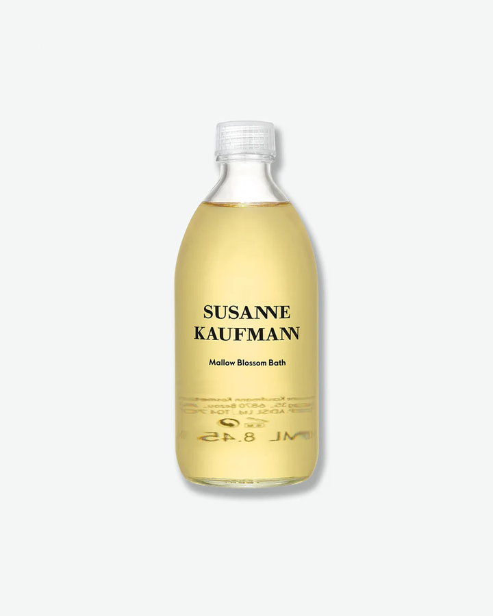 A product shot of a Susanne Kaufmann Mallow Blossom Bath bottle, centered against a plain light gray background. The curved clear glass bottle contains a translucent, pale golden liquid formula and is sealed with a simple, clear plastic ribbed screw cap. The front of the bottle features black text detailing the brand name and product title.