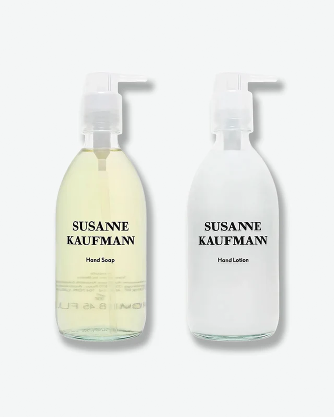 A product shot of two Susanne Kaufmann glass bottles arranged side-by-side against a plain light gray background. On the left is a clear glass bottle containing a translucent, pale yellow liquid formula labeled "Hand Soap". On the right is a frosted clear glass bottle filled with an opaque white formula labeled "Hand Lotion". Both are topped with identical clear plastic pump dispensers.