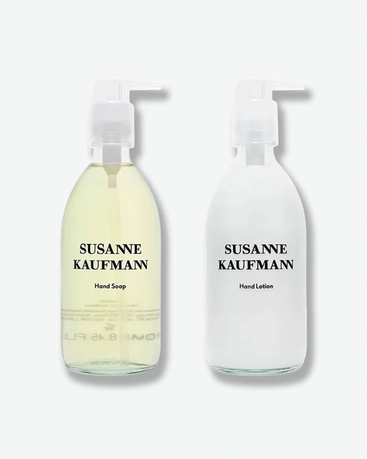 A product shot of two Susanne Kaufmann glass bottles arranged side-by-side against a plain light gray background. On the left is a clear glass bottle containing a translucent, pale yellow liquid formula labeled "Hand Soap". On the right is a frosted clear glass bottle filled with an opaque white formula labeled "Hand Lotion". Both are topped with identical clear plastic pump dispensers.