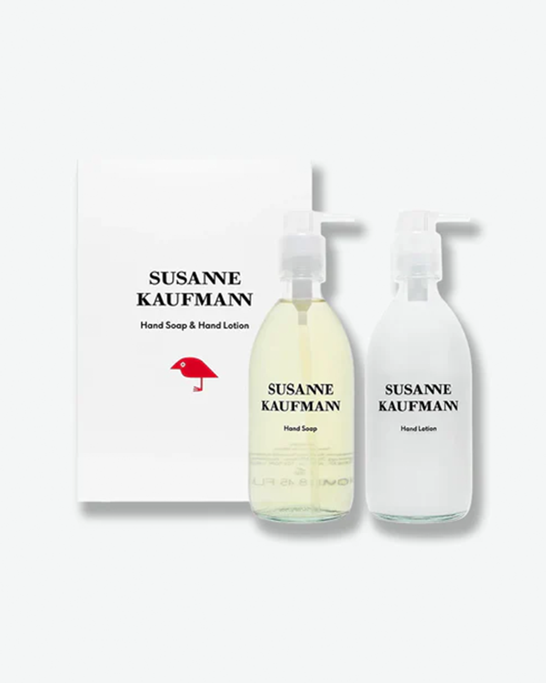 A product shot of a Susanne Kaufmann Hand Soap & Hand Lotion duo set standing in front of its white cardboard gift box, centered against a plain light gray background. The two clear glass pump bottles feature black text branding on the front. The white box behind them displays the brand text and a simple red illustration of a stylized bird.