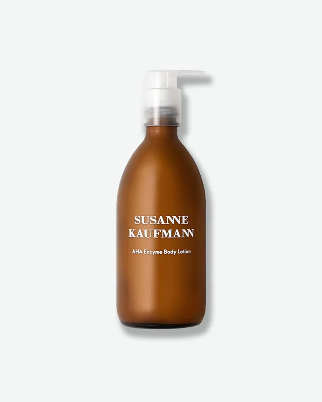 A product shot of a Susanne Kaufmann AHA Enzyme Body Lotion bottle, centered against a plain light gray background. The curved glass bottle features a semi-translucent, frosted amber-brown color and is equipped with a clear plastic pump dispenser. The brand name "SUSANNE KAUFMANN" is printed in serif capital letters in white on the front, above the product name.