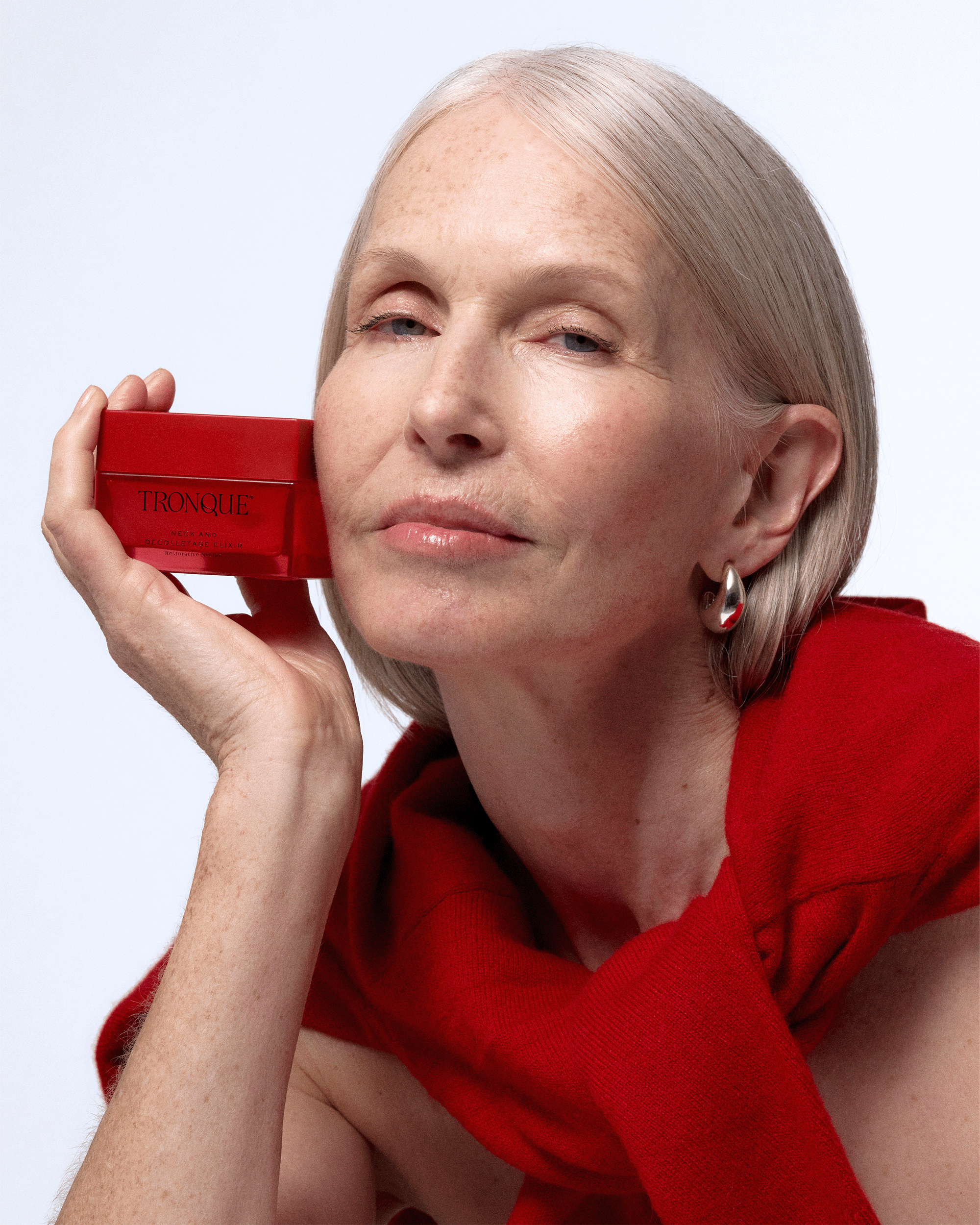Mature model with sleek gray hair holding a red Tronque Neck and Décolletage Elixir jar beside her face, wearing silver earrings and a red wrap.