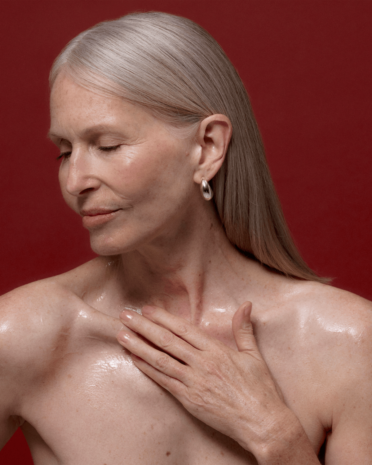 Mature model with long gray hair applying product across her neck and décolletage against a deep red background.