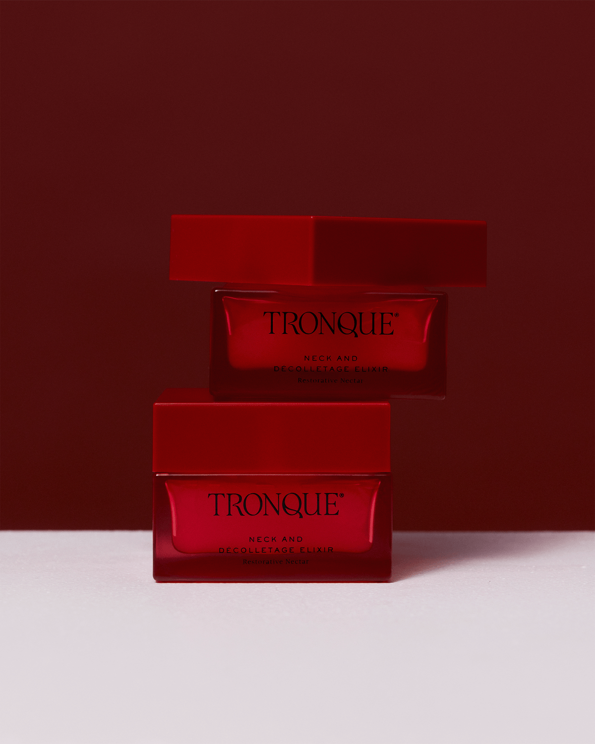Two red Tronque Neck and Décolletage Elixir jars stacked against a dramatic red backdrop with soft directional lighting.