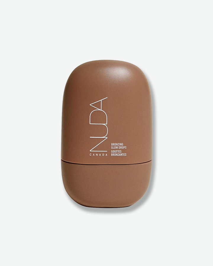 Front view of a NUDA Bronzing Glow Drops container centered against a plain gray background. The modern, pebble-shaped bottle has a matte, warm medium-brown finish and features the brand name "NUDA" printed vertically in a thin white font alongside the product details.