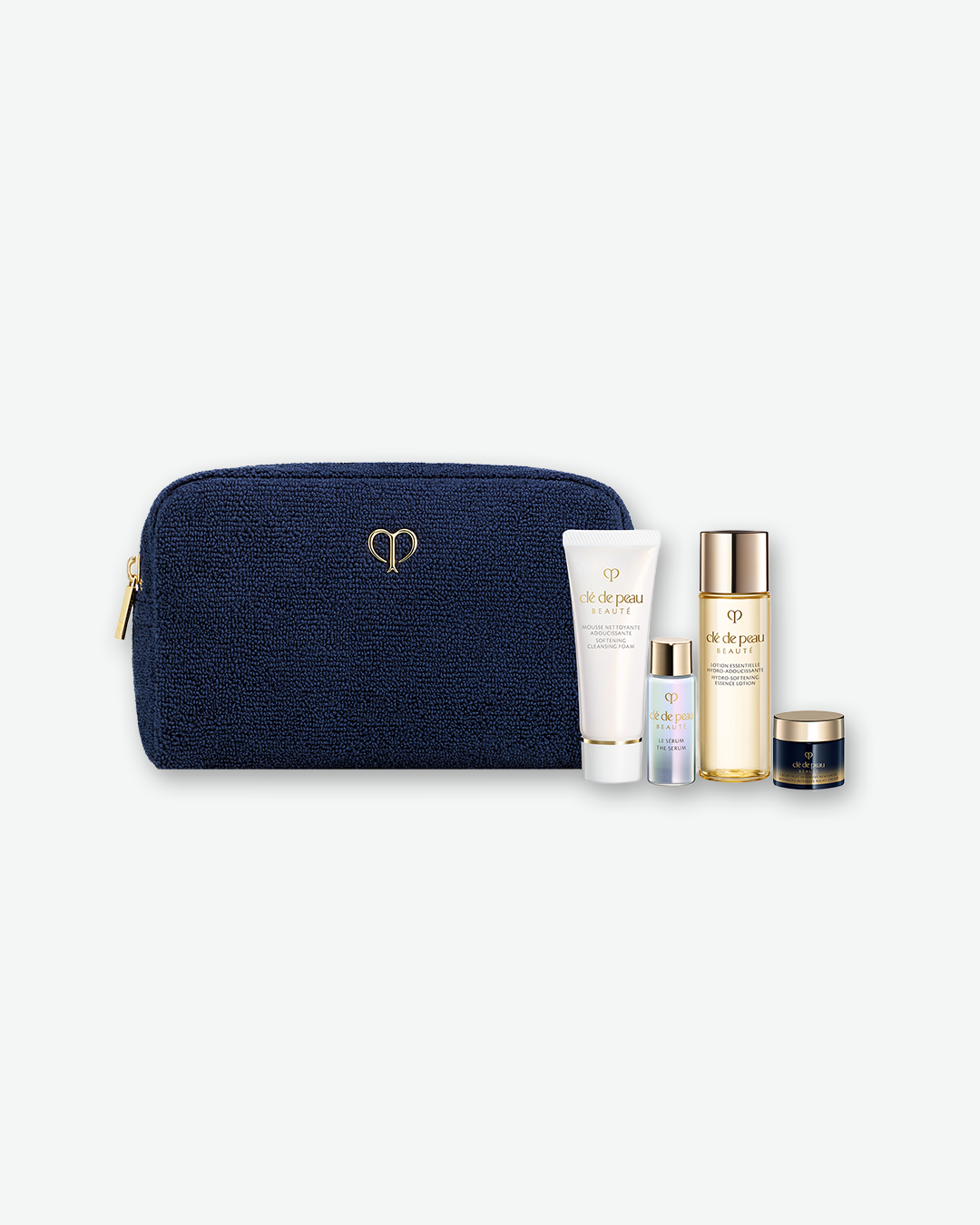 A Clé de Peau Beauté skincare travel set arranged neatly against a plain white background. On the left is a textured, dark navy blue fabric pouch featuring a small, golden logo in the center and a gold zipper pull. To the right of the pouch stands a collection of four miniature skincare products: a white tube of Softening Cleansing Foam, a small iridescent bottle of Le Sérum, a taller golden bottle of Hydro-Softening Essence Lotion, and a small dark blue jar of Intensive Night Cream.