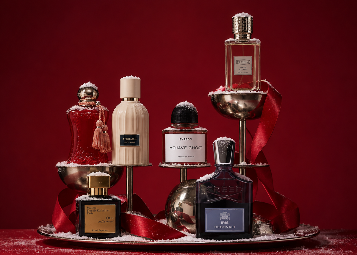 A curated collection of luxury perfume bottles arranged on a silver tray with red ribbons and white faux snow, set against a deep red background. Visible brands include Tom Ford, Jean Paul Gaultier, and Viktor & Rolf Spicebomb.