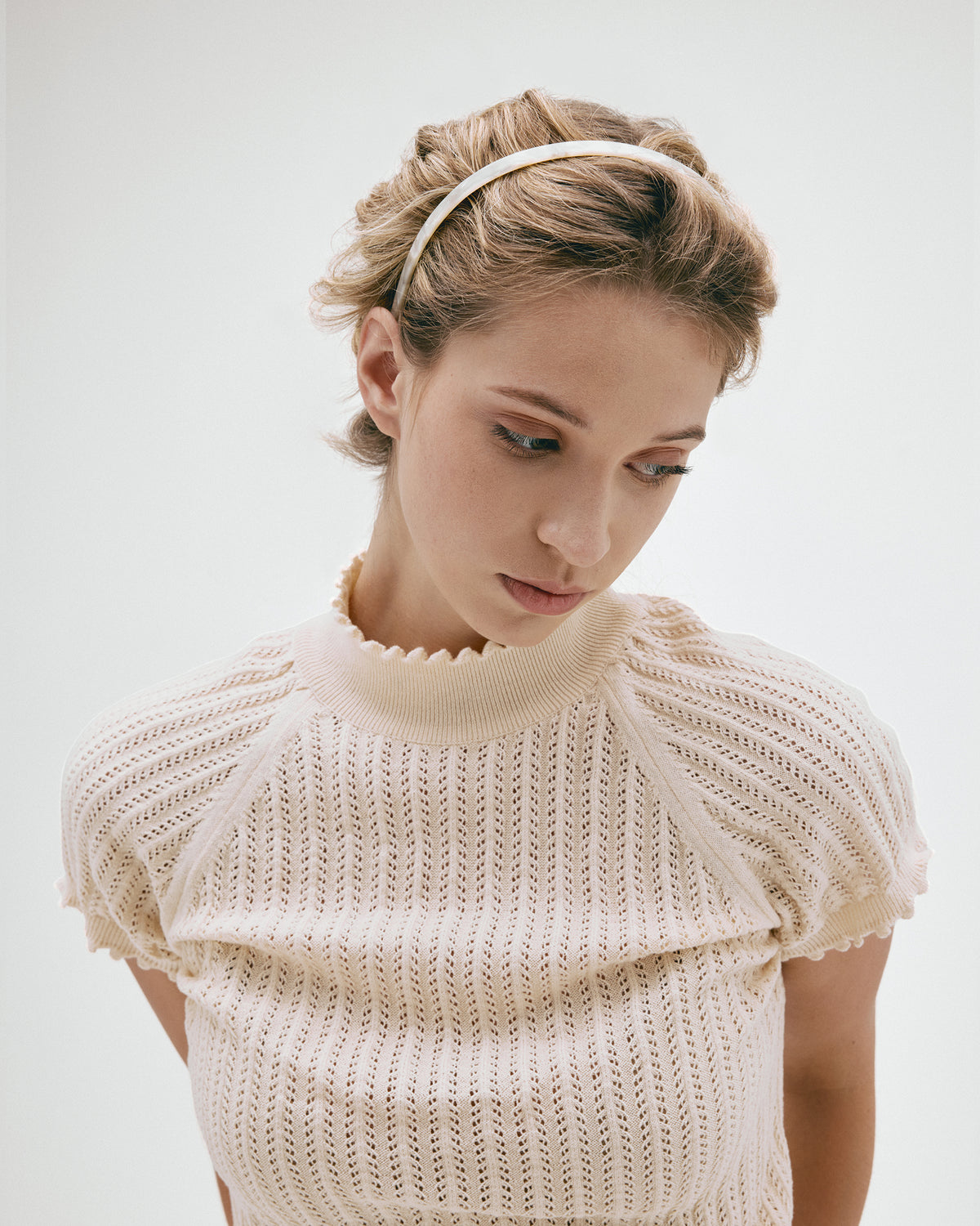 A woman wearing a cream-colored, short-sleeved knit top with a scalloped mock neck and a matching white headband, photographed against a plain light gray studio background.