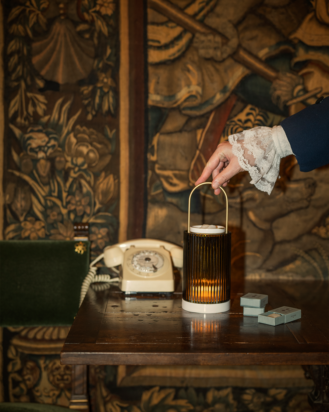 A hand in a lace-cuffed sleeve reaching to hold a brass-handled Trudon La Promeneuse wax warmer on a dark wooden table next to a vintage rotary phone and tapestry backdrop.