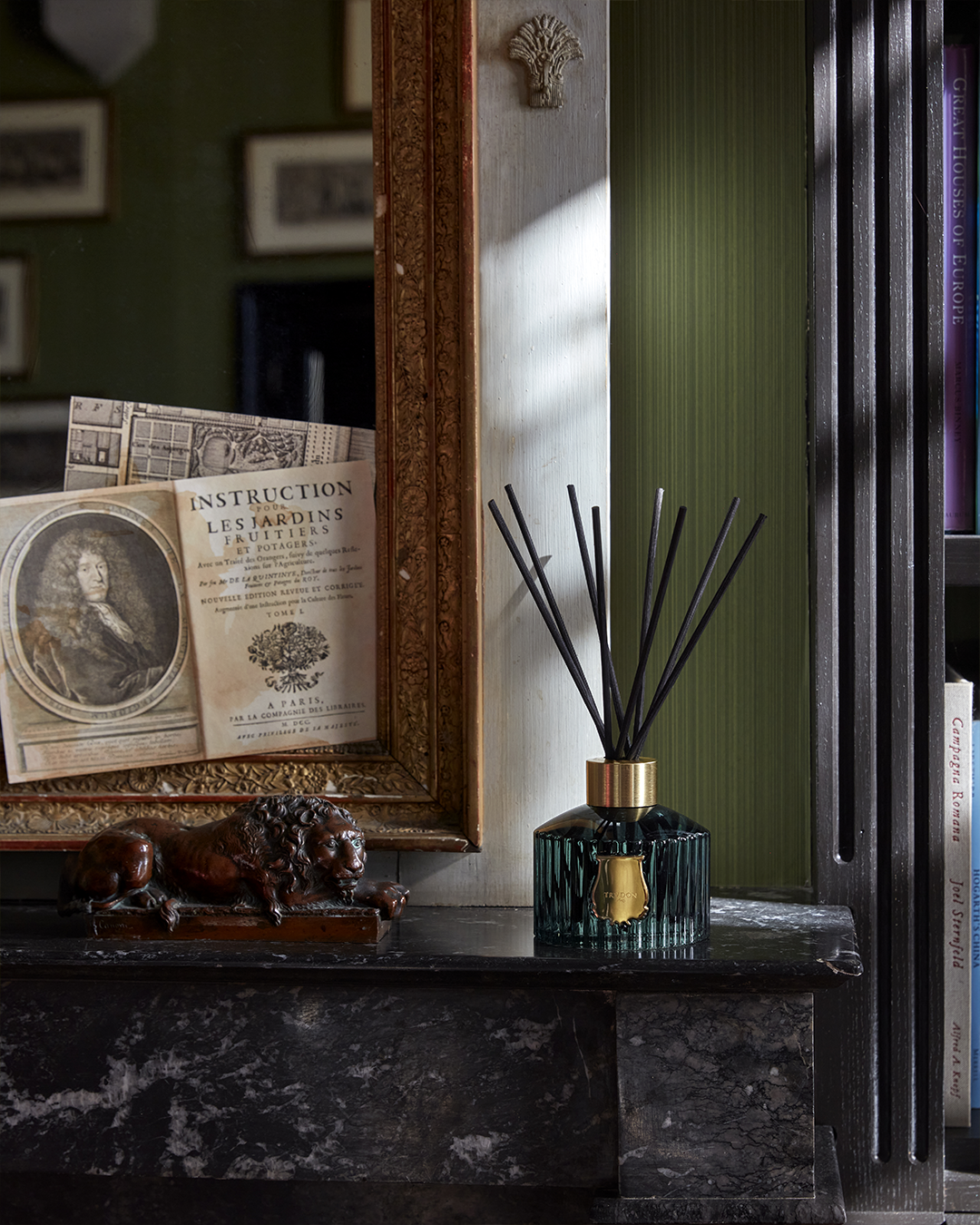 A Trudon reed diffuser sitting on a dark marble fireplace mantel next to a small bronze lion statue and framed art.