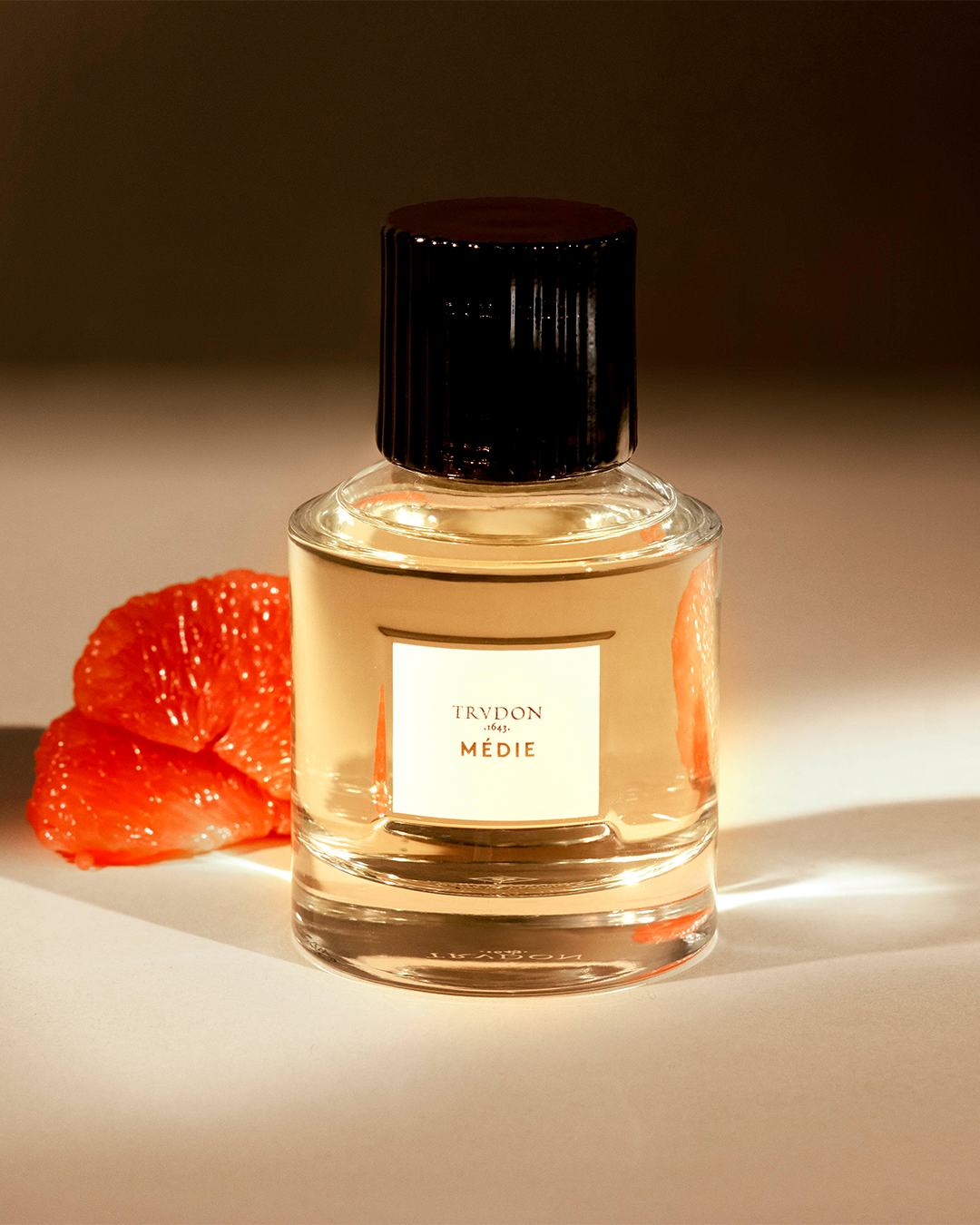 A glass bottle of Trudon Médie perfume with a black cap, standing next to a slice of fresh grapefruit under warm, dramatic lighting.