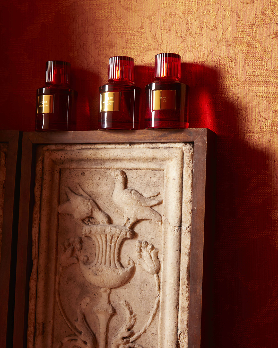 Three red glass Trudon perfume bottles aligned on top of a carved stone wall relief against a patterned orange wallpaper background.