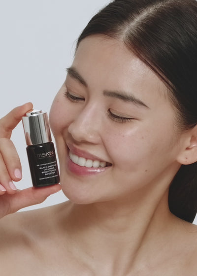 A smiling model holds a small dark bottle of 111SKIN serum beside her face. The video demonstrates how to use the treatment, showing the product being shaken before application and applied to clean skin as part of a skincare routine. Text on screen instructs users to shake well before use.