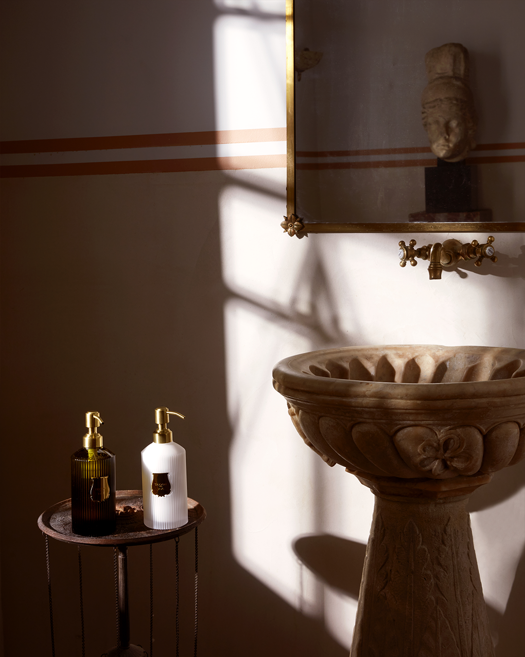 Two ribbed Trudon pump bottles—one dark amber glass and one white—sitting on a small side table next to a carved stone pedestal sink illuminated by soft sunlight.